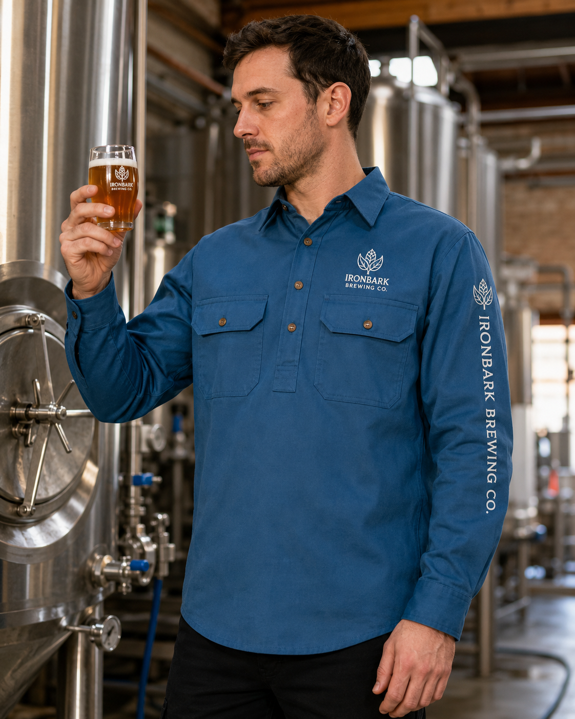 A man in a blue long-sleeve Ironbark Brewing Co. button-down shirt stands in a brewery, holding up a glass of amber beer.