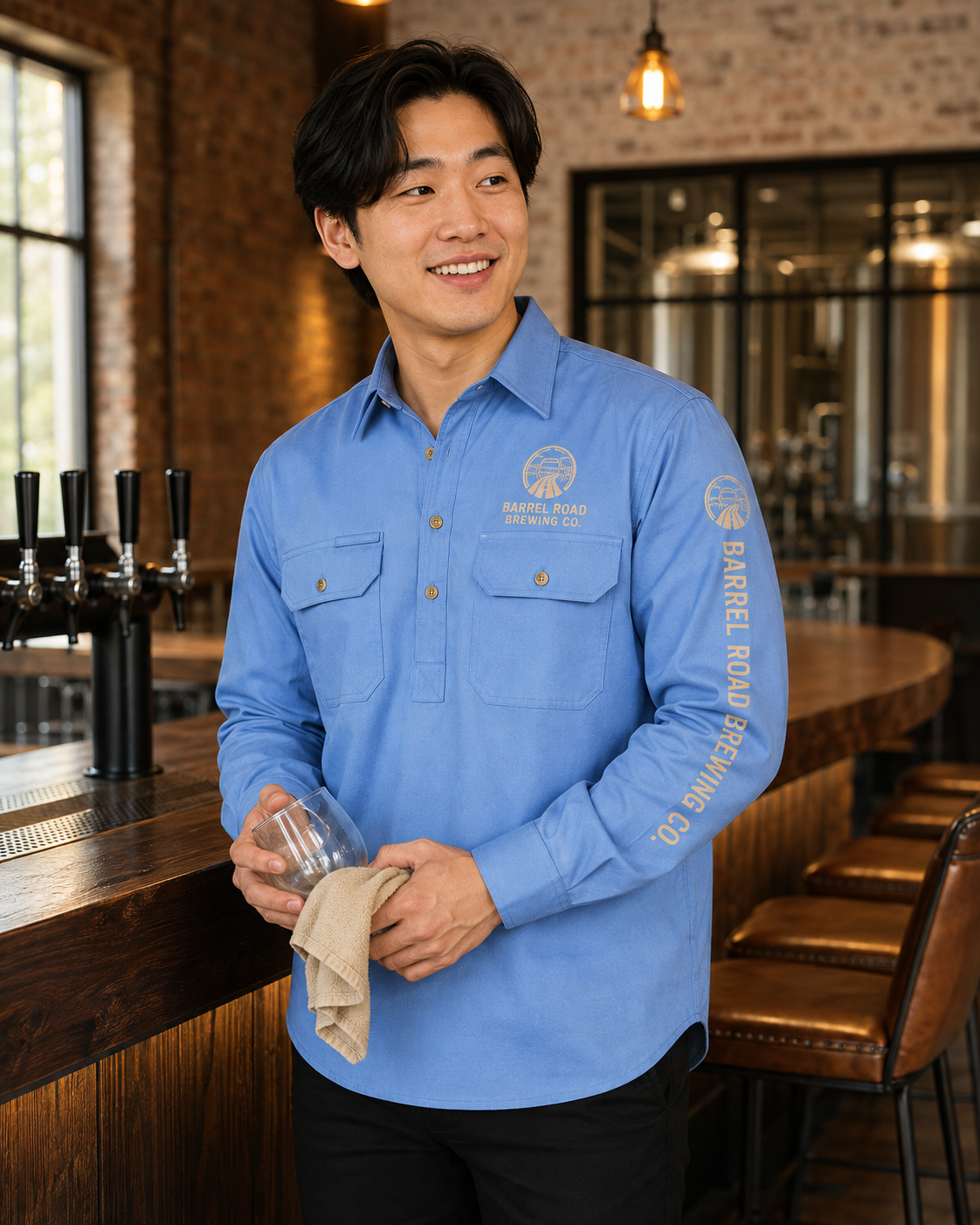 A man wearing a light blue long-sleeve button-up shirt with 'Falcon Barrel Brewing Co.' printed on the chest and sleeve, standing in a bar environment and holding a glass and a cloth.