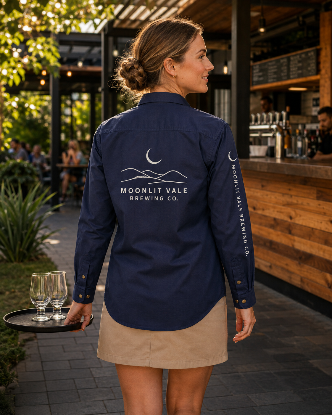 A woman standing outdoors at a brewery bar, shown from the back wearing a navy long-sleeve button-up shirt with white Moonlit Vale Brewing Co. logos on the back and sleeve, paired with a beige mini skirt and holding a tray with wine glasses.
