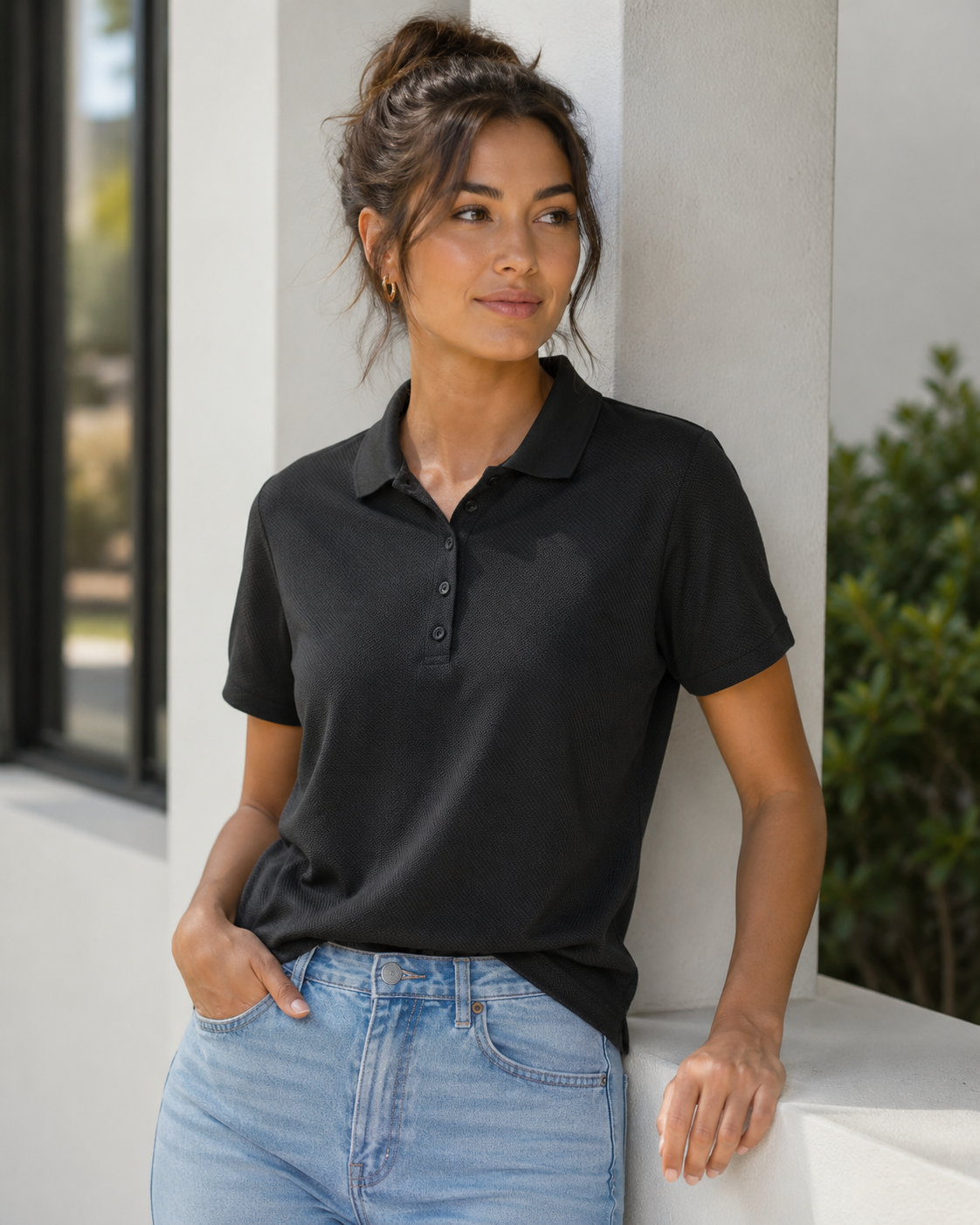 A woman wearing a black short-sleeve polo shirt and light blue jeans, leaning against a white architectural column in a bright, modern setting.