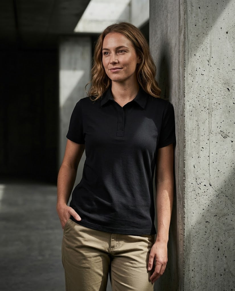 A woman wearing a black short-sleeve polo shirt and khaki pants, leaning against a concrete pillar in a modern architectural setting.