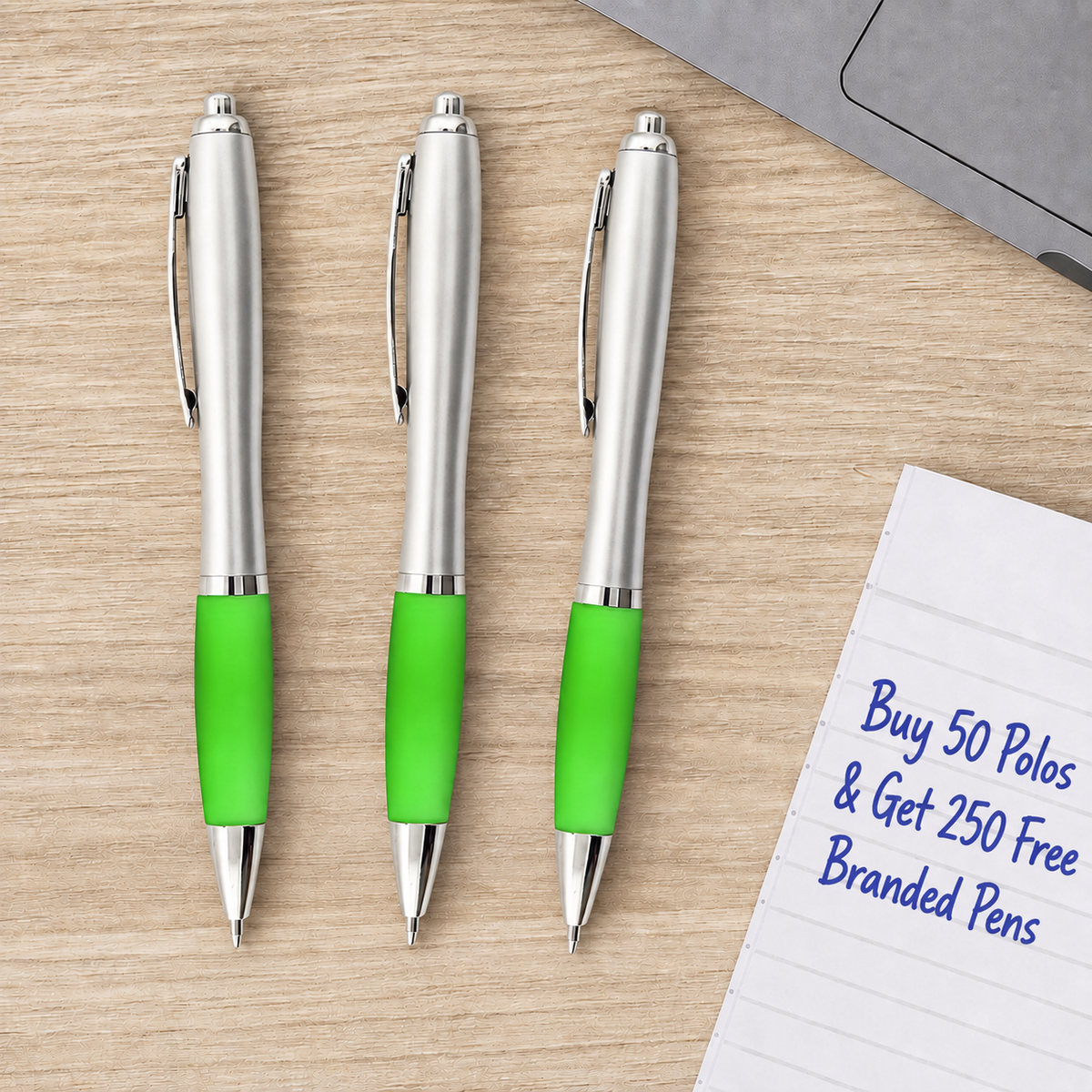 Three silver retractable pens with bright green grips arranged on a light wood surface next to a notepad that reads 'Buy 50 Polos & Get 250 Free Branded Pens'.