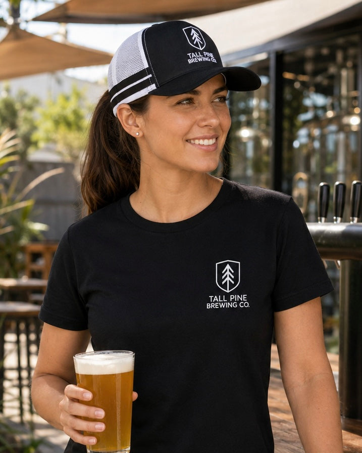 A woman wearing a black and white trucker hat and a black t-shirt with a white Tall Pine Brewing Co. logo, holding a glass of beer at an outdoor bar.