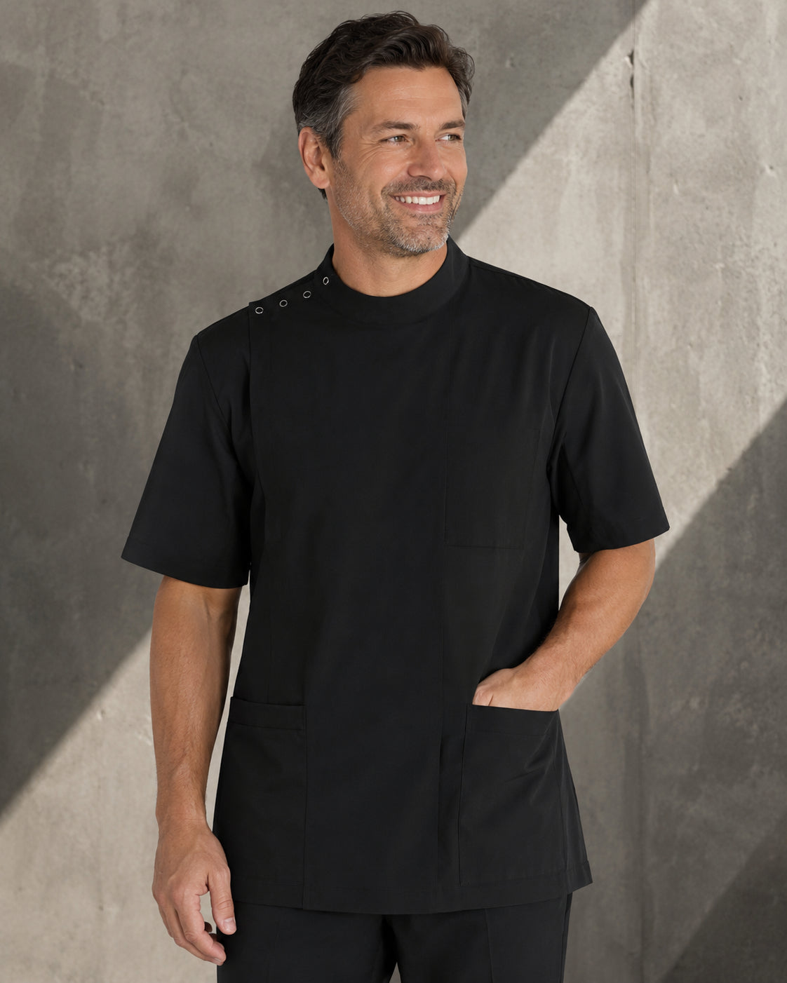 A man wearing a black short-sleeve scrub top with three decorative buttons on the shoulder and front pockets, standing against a textured gray wall.