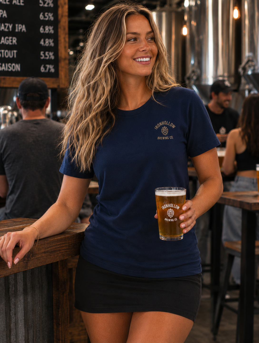 A woman smiling in a brewery setting, wearing a navy blue crewneck t-shirt with a gold 'Brookfield Brewing Co. Avonlea Ohio' logo on the left chest, and holding a pint of beer.