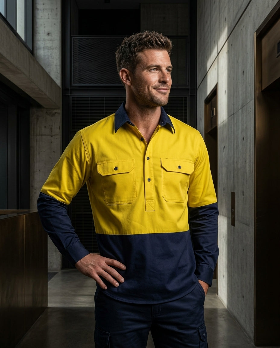 A man wearing a two-tone long-sleeve work shirt in bright yellow and navy blue, standing in a modern concrete-walled corridor.