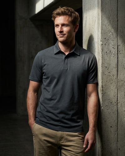 A man wearing a dark charcoal gray short-sleeve polo shirt and khaki pants, leaning against a textured concrete wall in a dimly lit industrial setting.