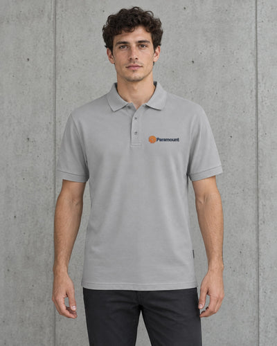 A man wearing a light gray short-sleeve polo shirt with a small orange and black Paramount logo on the left chest, standing against a textured gray concrete wall.