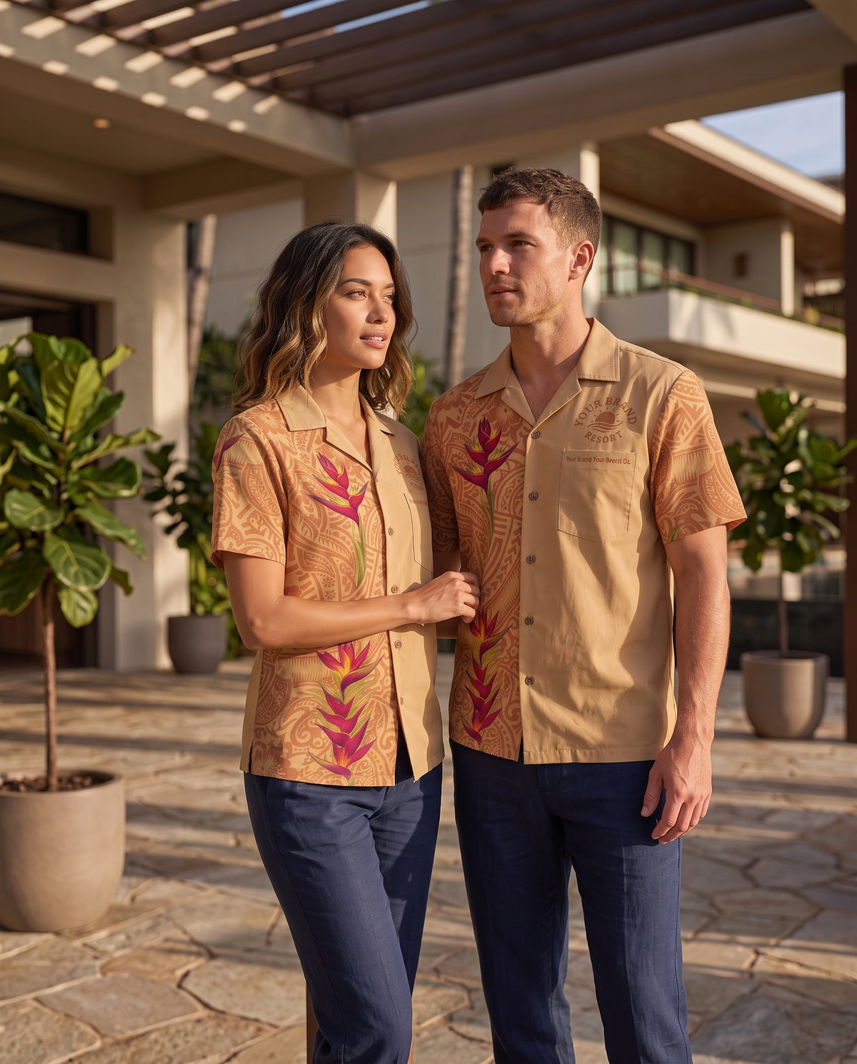 A man and a woman standing outdoors in front of a modern house, wearing matching tan short-sleeve camp-collar shirts with a tropical floral print in pink and green, paired with navy pants.