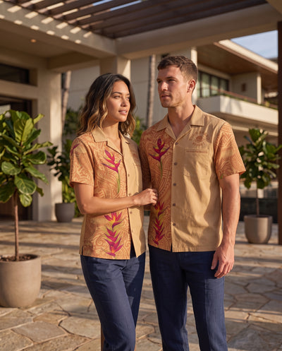 A man and a woman standing outdoors in front of a modern house, wearing matching tan short-sleeve camp-collar shirts with a tropical floral print in pink and green, paired with navy pants.